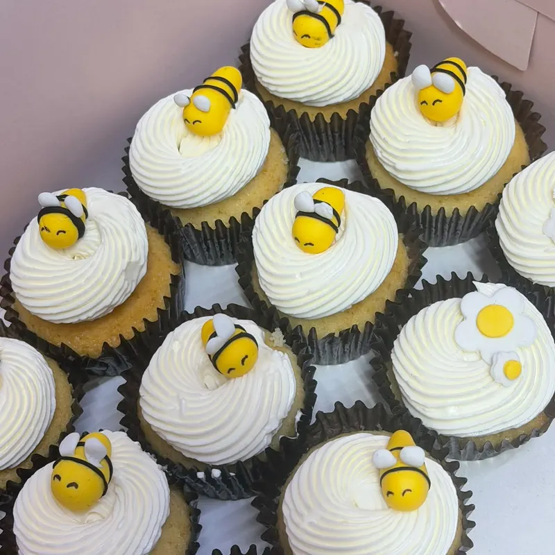 A box of vanilla cupcakes with swirled cream and hand-shaped bee toppers